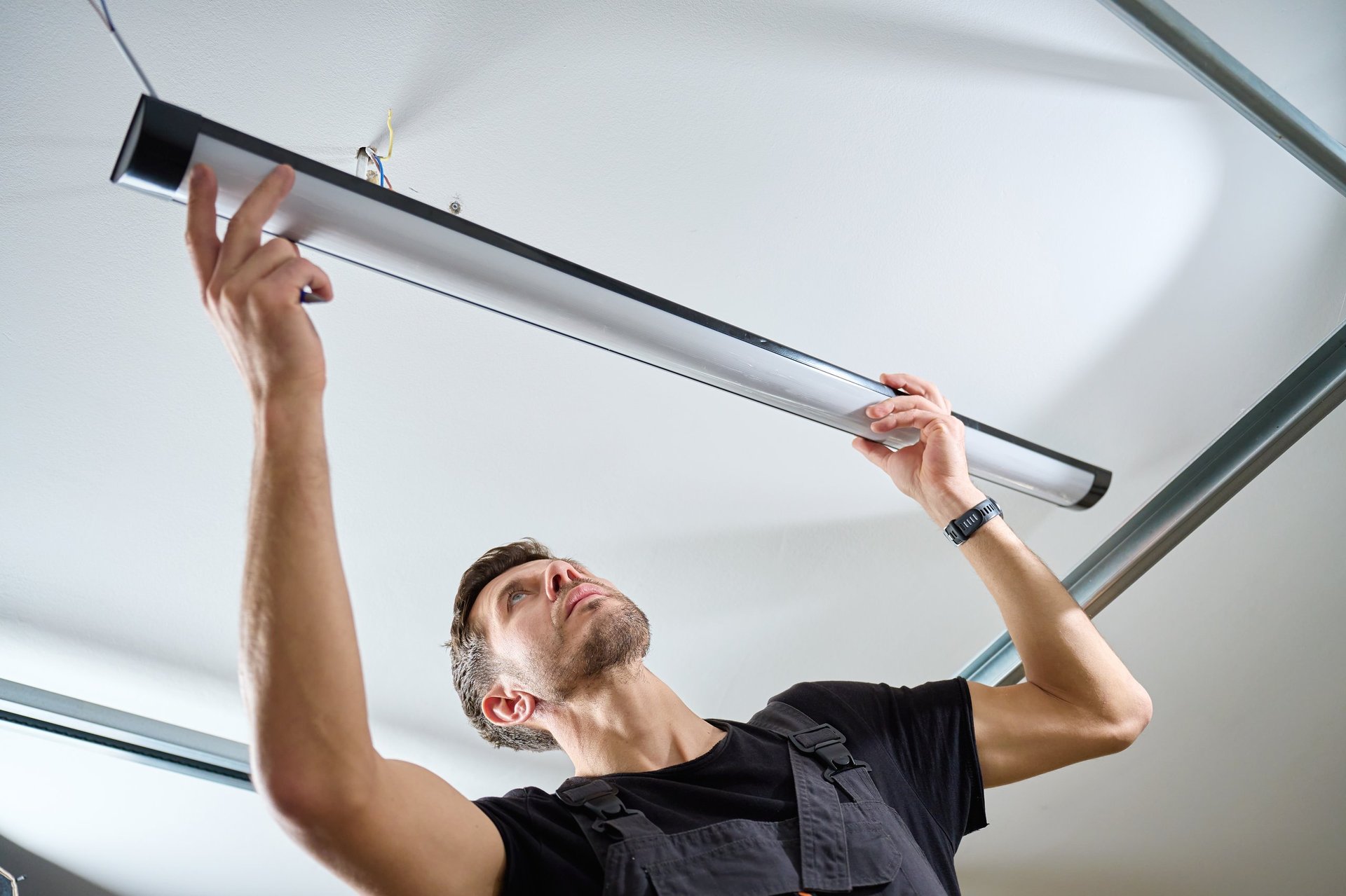 Man in workwear installing a linear LED light fixture on the ceiling indoors. Concept of electrical installation, maintenance work and home renovation.