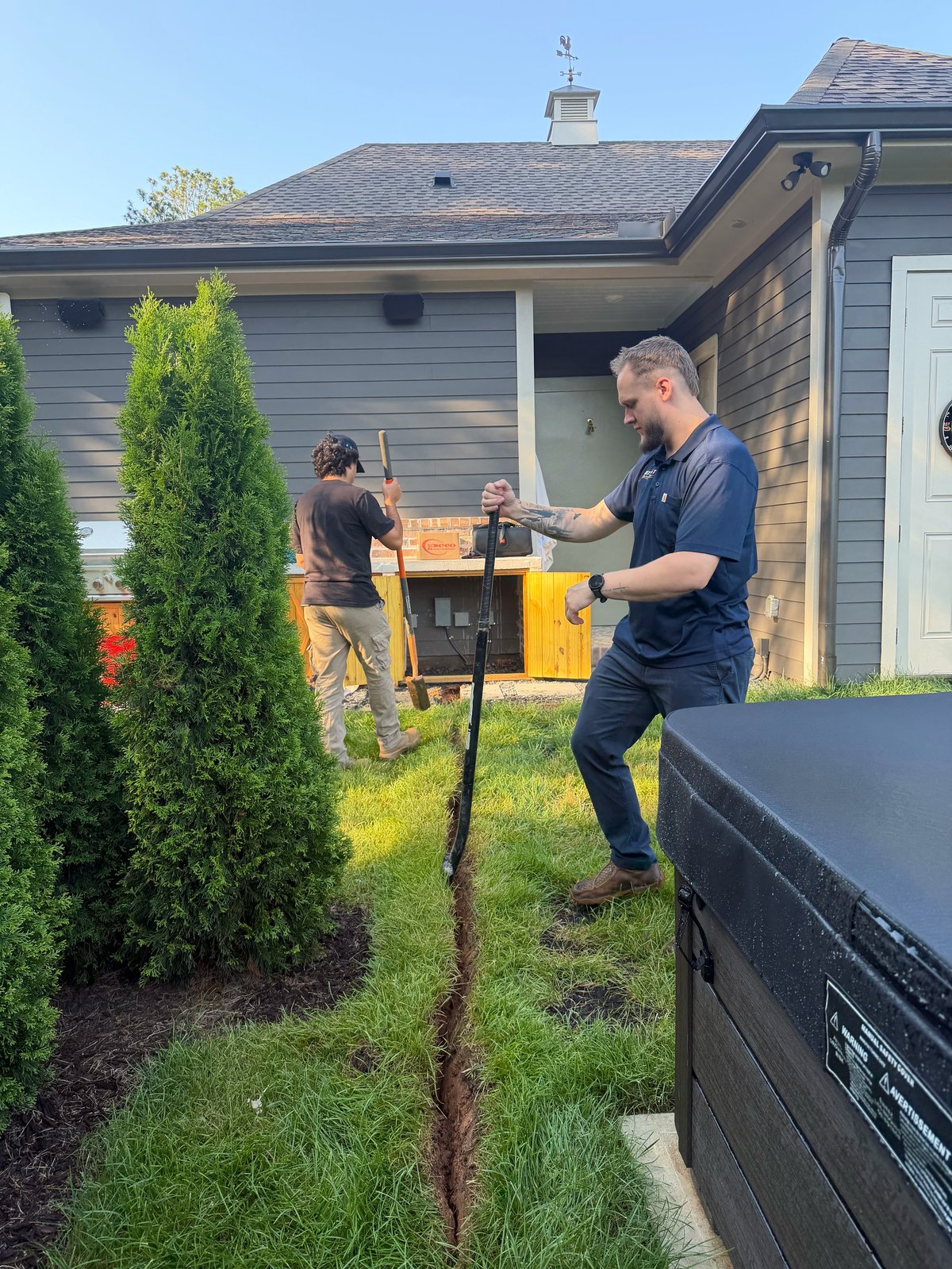 Two people grilling food on a yellow outdoor grill in a backyard next to a house and evergreen shrub