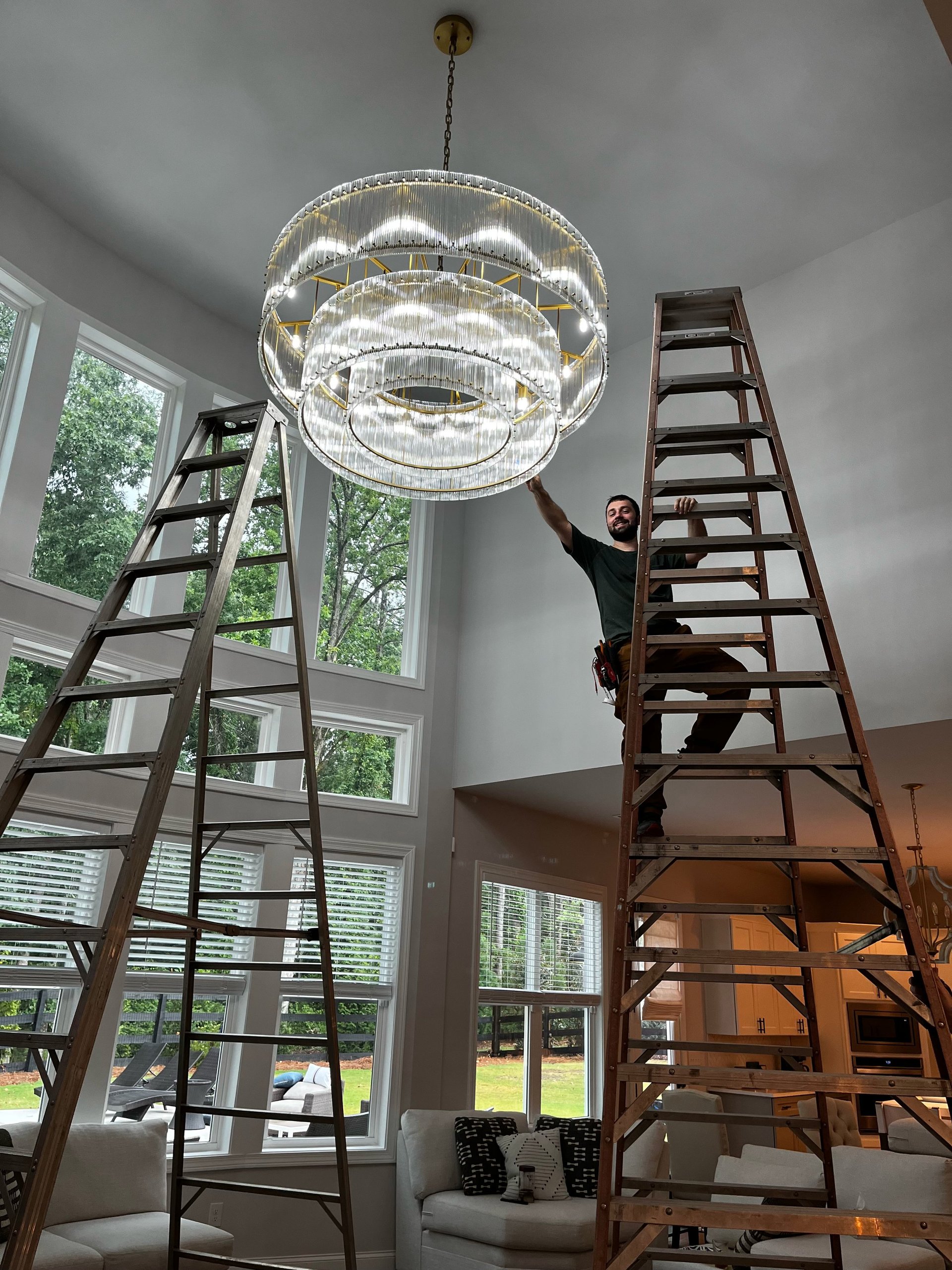 Person on tall wooden ladder installing or adjusting a modern circular crystal pendant light fixture in a bright room with large windows