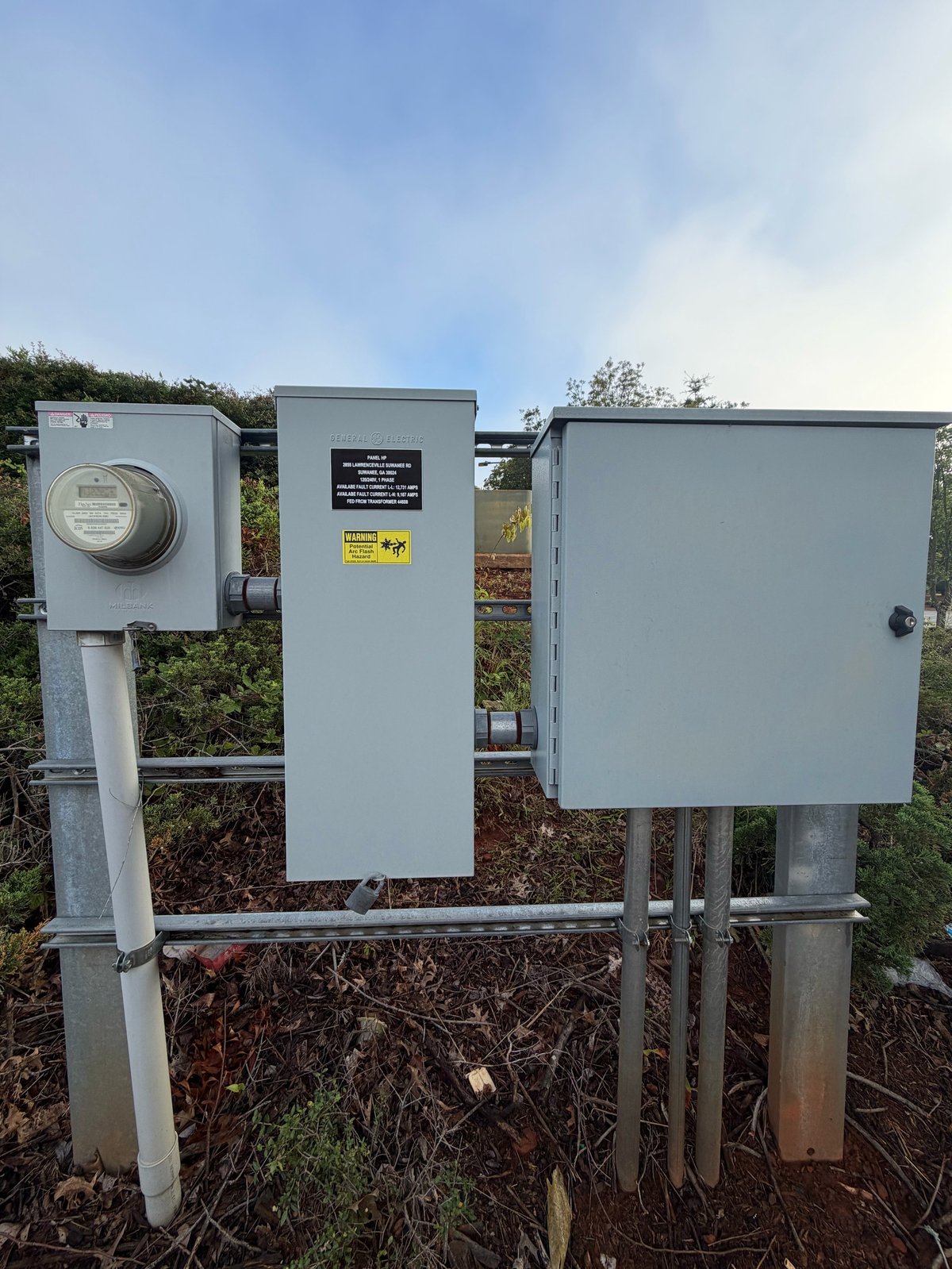 Three gray electrical utility boxes mounted on a metal pole in an outdoor area with vegetation and sky in background