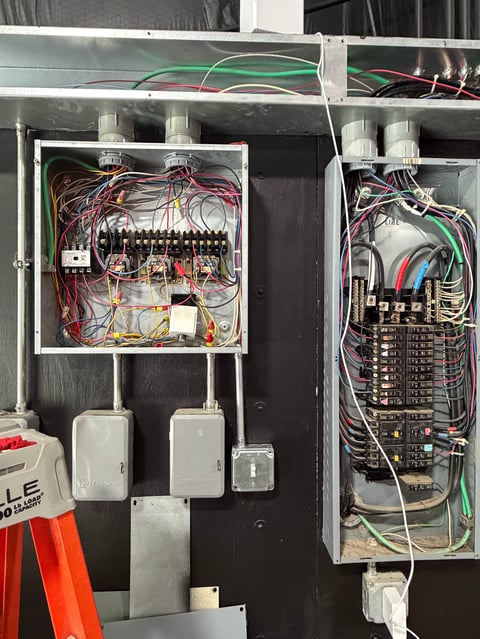 Electrical panel installation showing two circuit breaker boxes with colored wiring, one with terminal strips on left and main breaker panel on right, mounted on black wall.