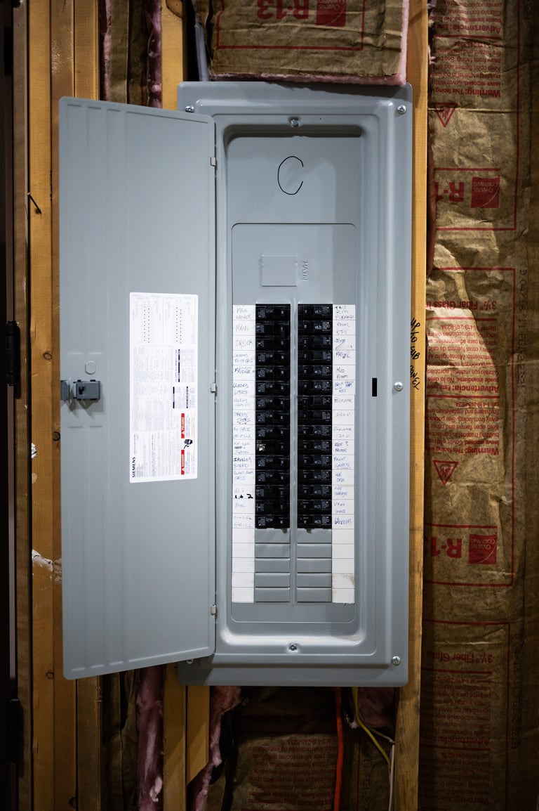 Gray electrical panel box mounted on wooden wall with open door showing circuit breakers and documentation label inside