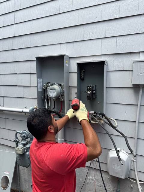 Electrician in red shirt and safety gear installing or servicing electrical panel and meters on white siding house exterior