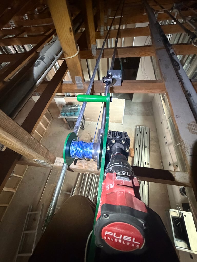 Overhead view of construction equipment setup showing a red power drill with blue wire spool mounted on green frame suspended in wooden attic structure
