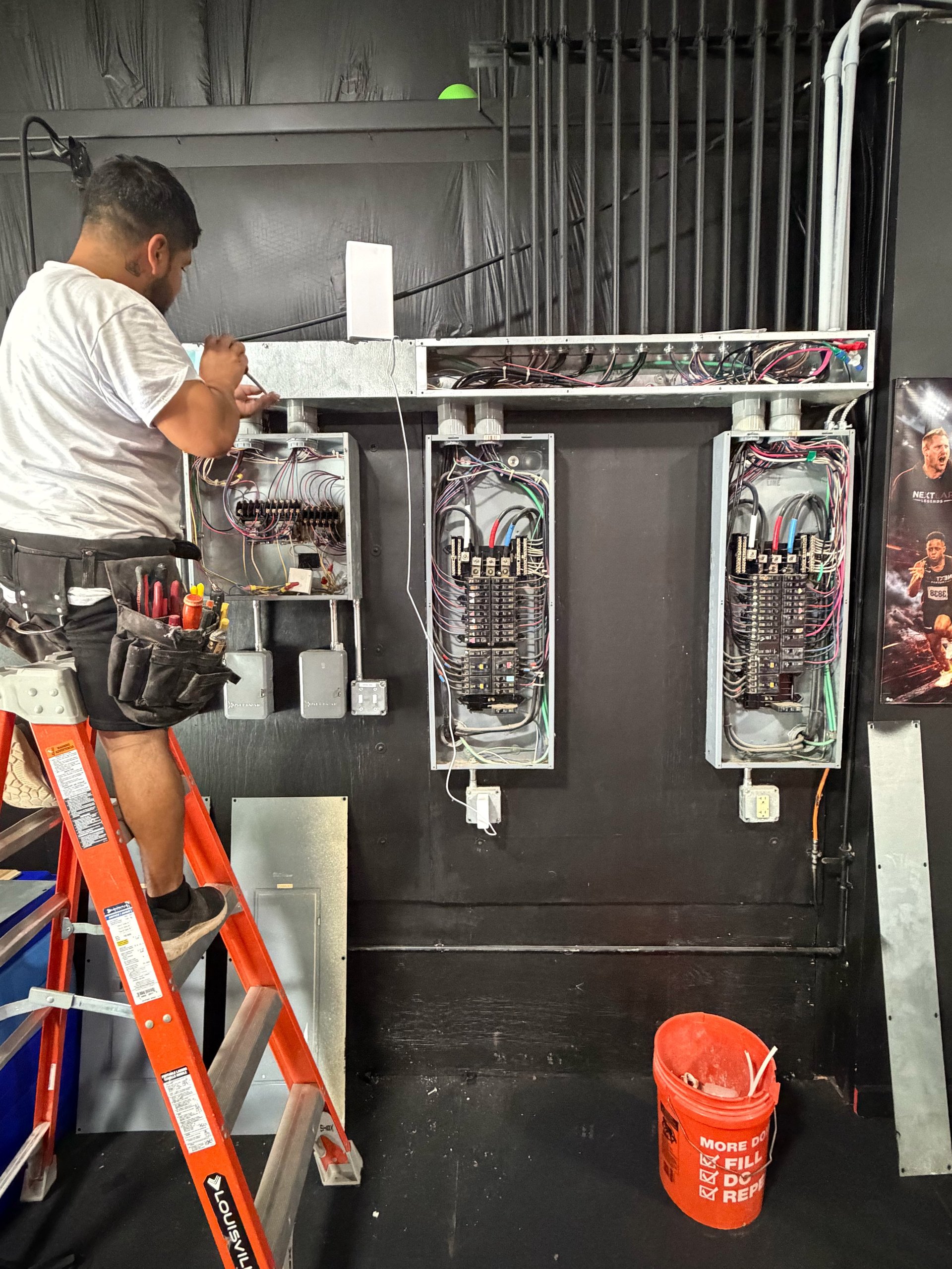Electrician on ladder working on electrical panel with exposed wiring and breakers