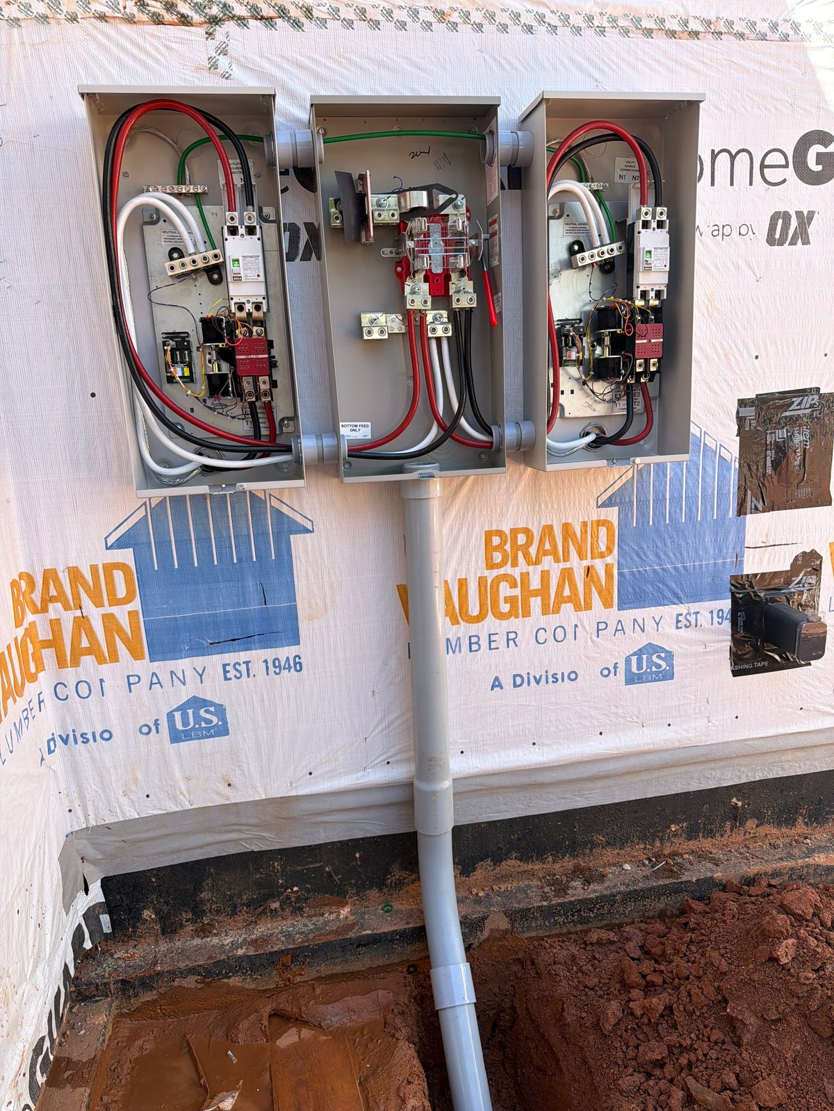 Three electrical service panels mounted on a white outdoor utility box labeled Brand Vaughan, with red and white wiring, installed above muddy ground