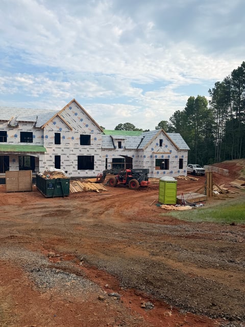 Under-construction residential house with white exterior, exposed framing, construction equipment, and red clay site on sunny day