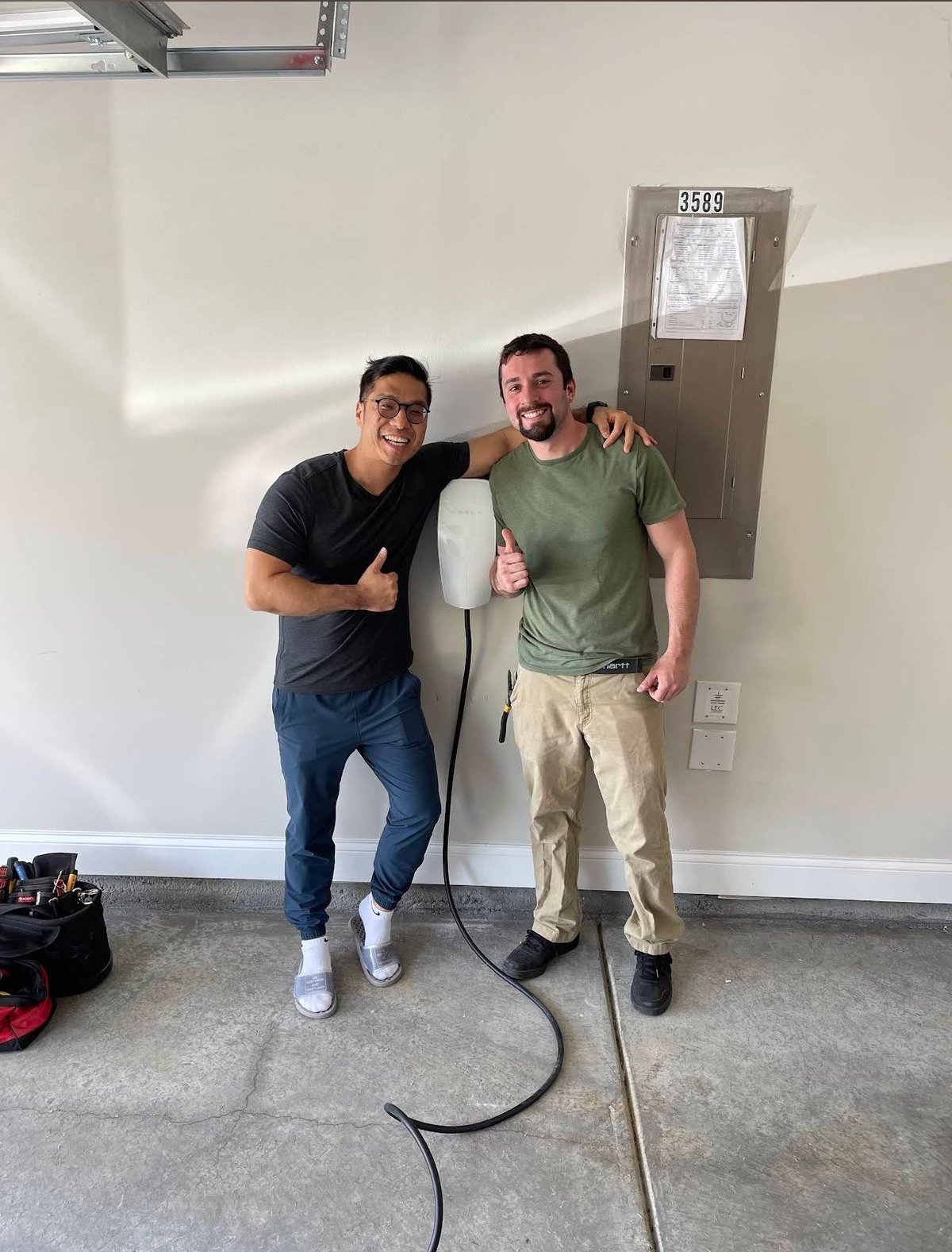 Two men posing together indoors with arms around each other, standing in front of a white wall with an electrical outlet and door marked 3858.