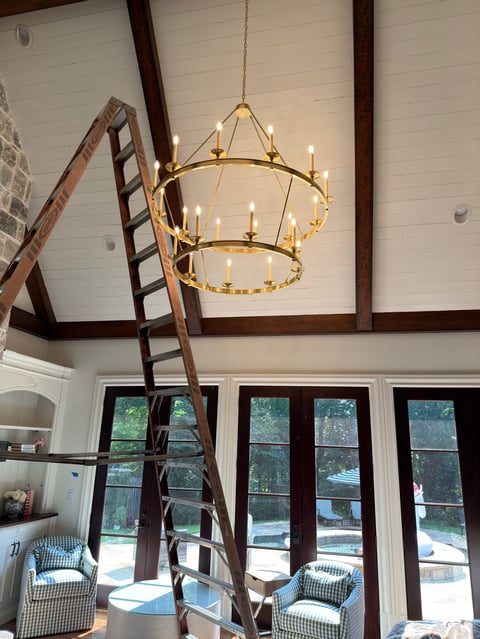 Gold chandelier with lit candles hanging in high-ceilinged room with wooden beams and ladder leaning against wall