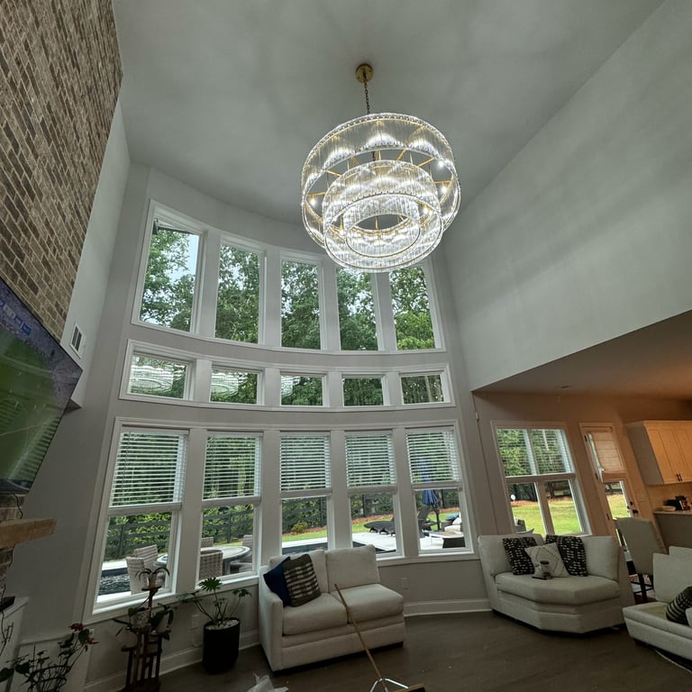 Modern living room with two-story windows, contemporary glass pendant light, and cream-colored furniture overlooking green landscape