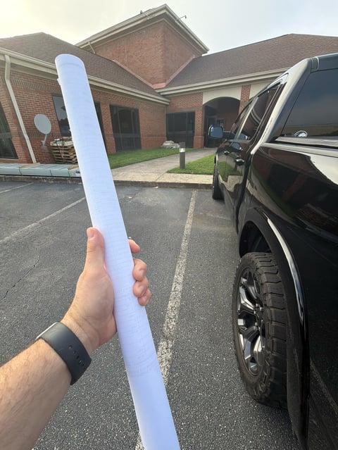 Persons hand holding a white foam pool noodle in a parking lot with a brick building and black SUV in the background