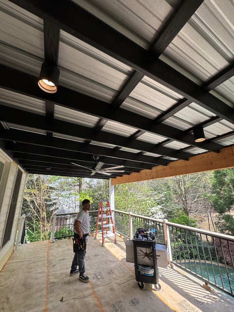 Covered deck with black pergola and polycarbonate roof panels, person standing on wooden floor with railing and wooded landscape view