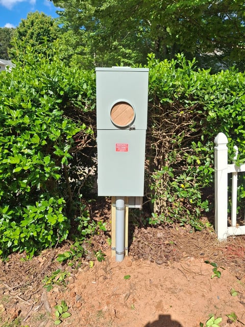 White rectangular utility box mounted on a metal post in a garden surrounded by green shrubs and trees