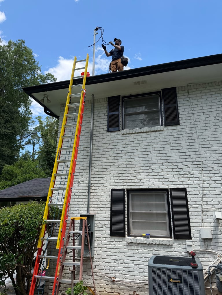 Worker on tall yellow ladder cleaning gutters on white brick two-story house roof on sunny day