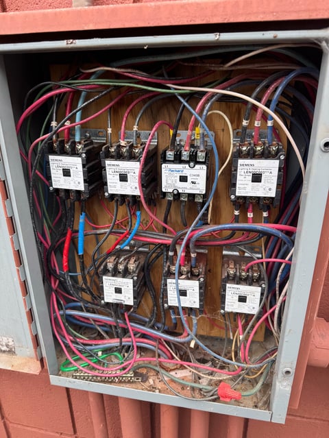 Electrical panel with multiple contactor switches, colorful wiring, and labeled terminals mounted on a wooden backing inside a metal enclosure