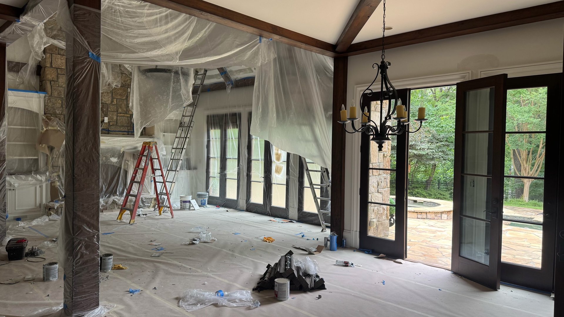 Interior construction site with plastic sheeting, red ladder, and glass doors leading to outdoor patio area