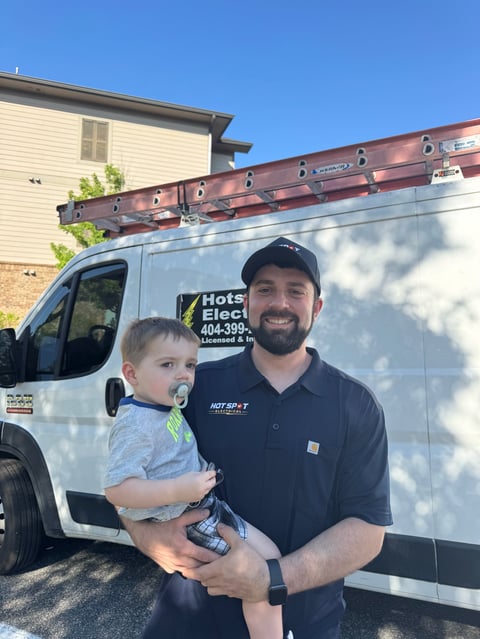 Smiling electrician holding a young child in front of a white service van with ladder rack parked at a residential property