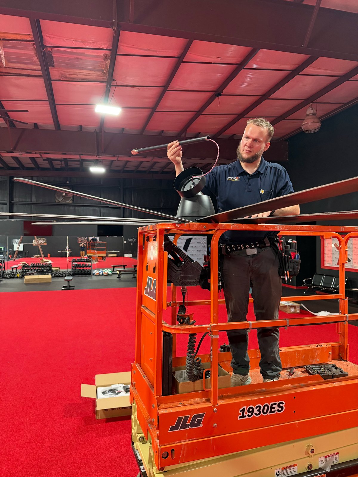 Man standing on an orange JLG aerial lift platform holding up a helmet in an indoor warehouse with red flooring and ceiling lights.