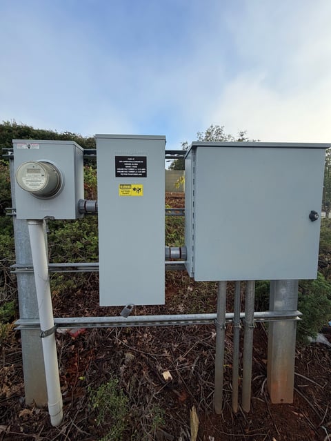 Three gray electrical utility boxes mounted on metal posts in an outdoor setting with vegetation