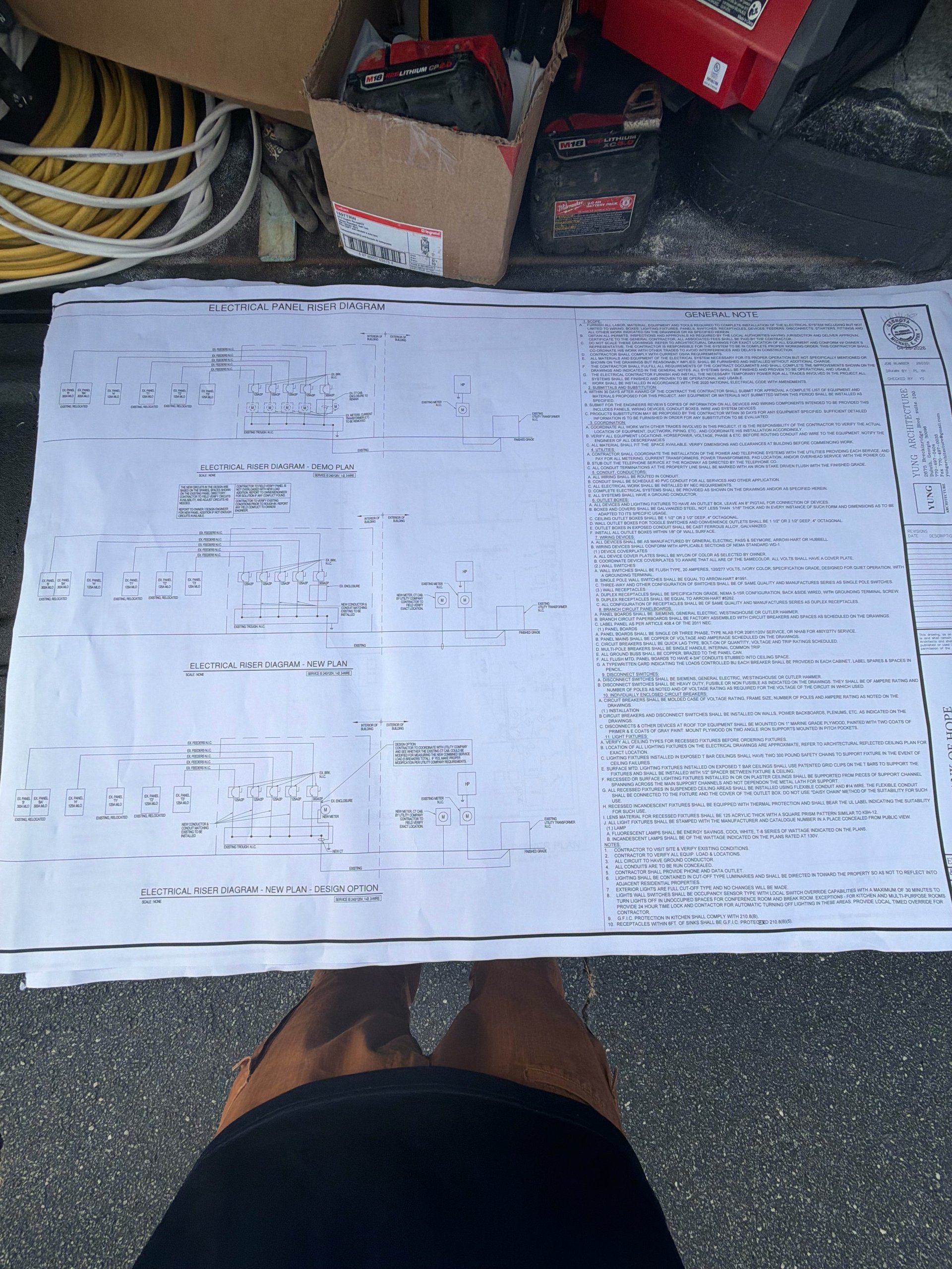 Electrical schematic blueprints spread on floor with equipment boxes and coiled cable visible in background