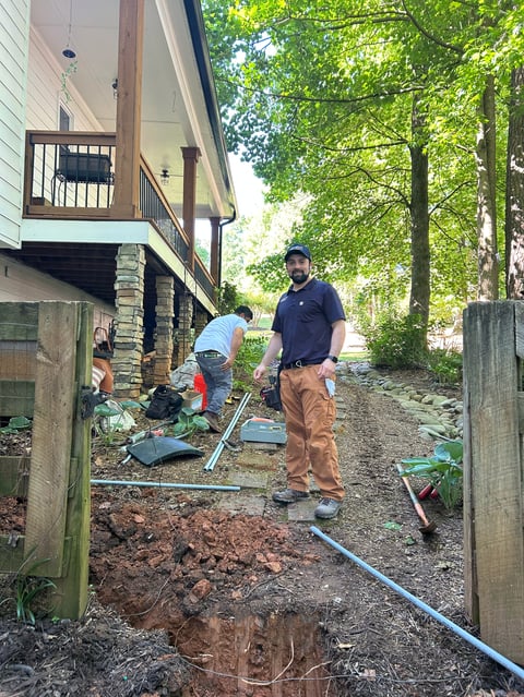 Two workers performing yard excavation near a house, with exposed soil and equipment visible among trees