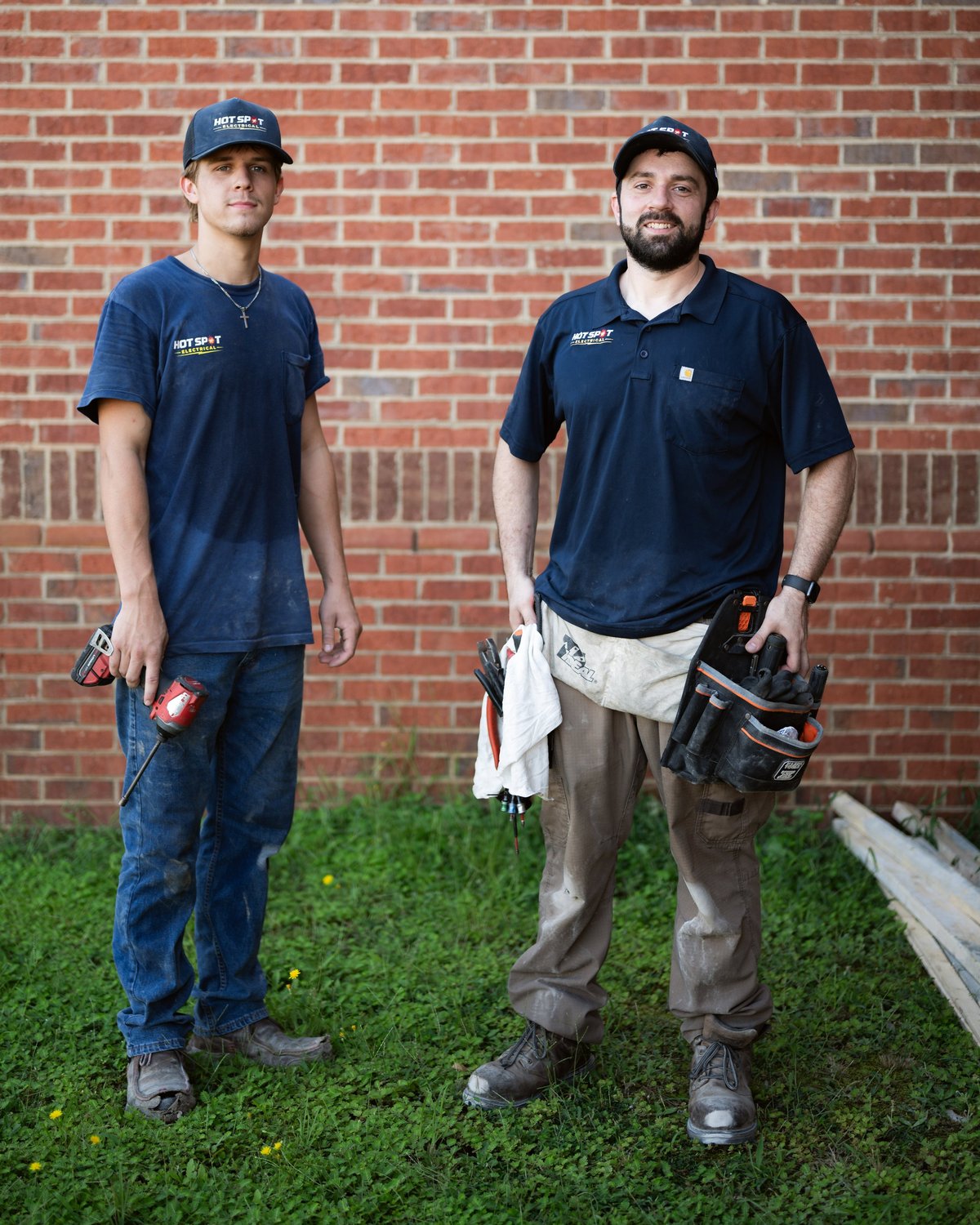 Two tradesmen in navy work shirts and caps stand against a brick wall, holding tools and wearing tool belts