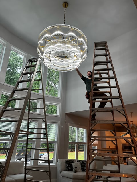Person on ladder installing a large modern crystal chandelier hanging from ceiling in bright room with tall windows