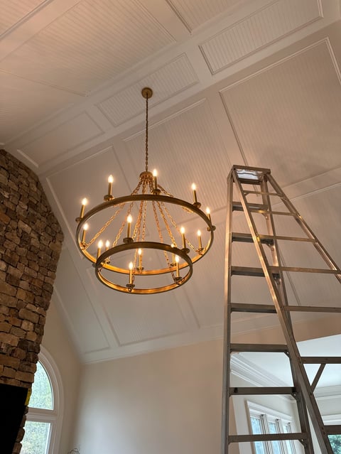 Gold circular chandelier with lit candles hanging in a white coffered ceiling room with a ladder and brick wall visible