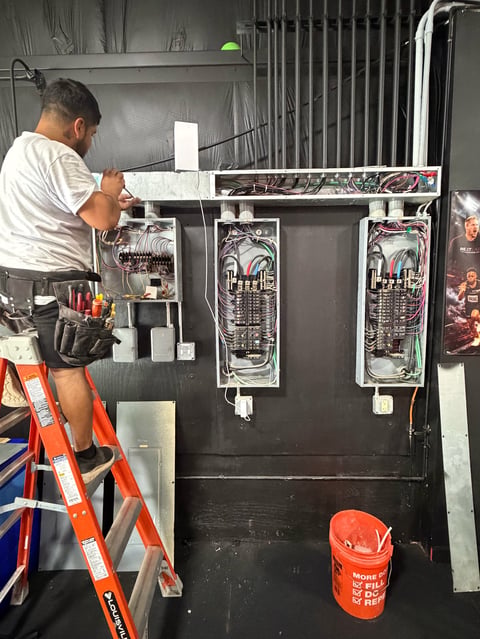 Electrician on ladder working on interior electrical panel with exposed wiring and circuit breakers in an unfinished installation space