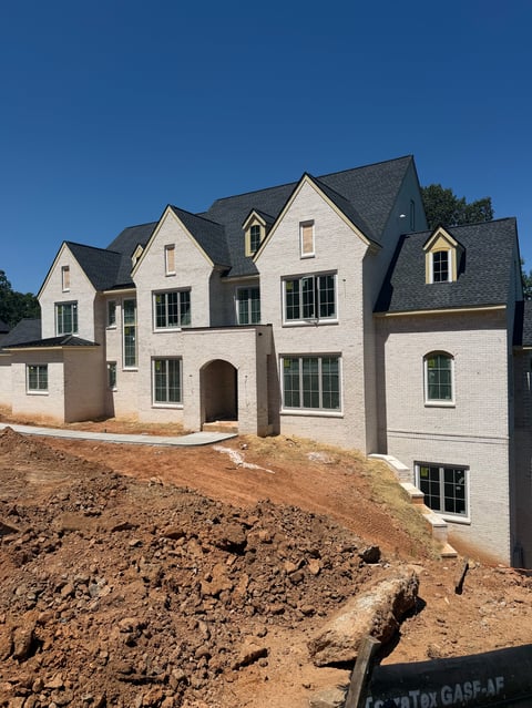 Modern white brick house under construction with dark gray roof on excavated lot against blue sky