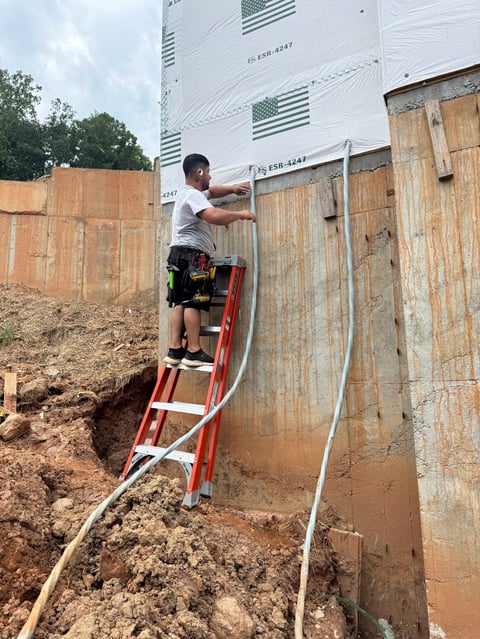 Worker on red ladder installing electrical conduit on RV exterior with dirt ground and fencing visible