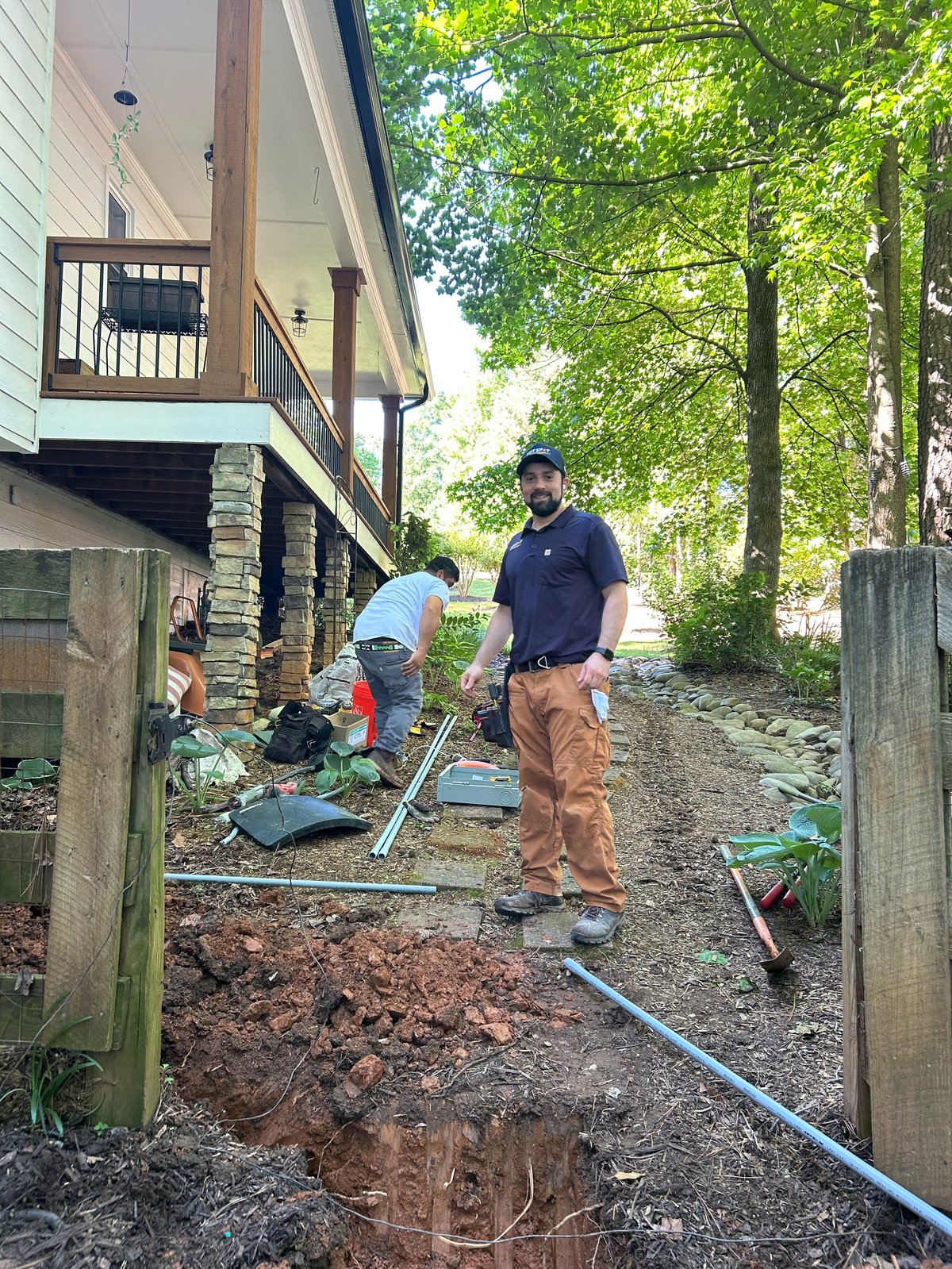 Two workers performing yard excavation and landscaping work next to a residential house with exposed soil and equipment visible