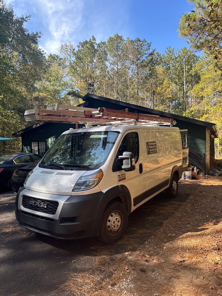 Beige van with extended roof rack and awning parked on dirt ground under tall trees in daylight